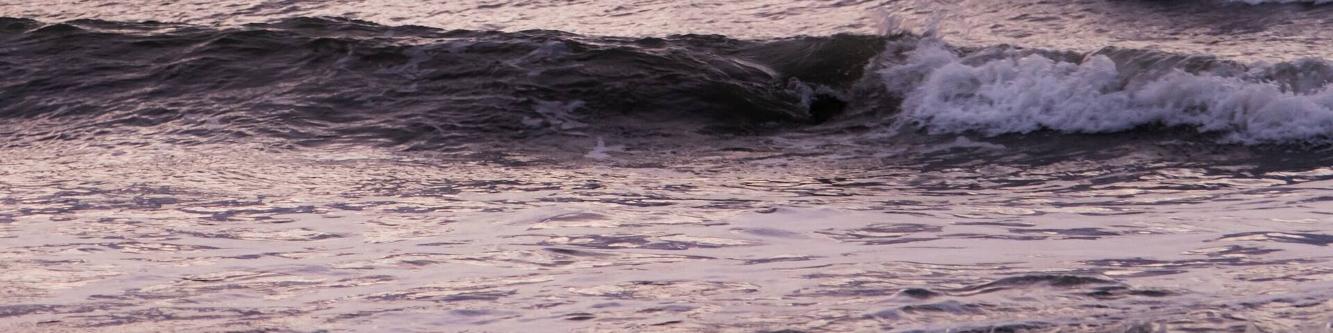 A pattern of three waves making their way to the beach and crashing on the shallow water. Each wave has a whitecap. The ocean is beneath a cloud filled sky lit by the rising sun with pink and orange.