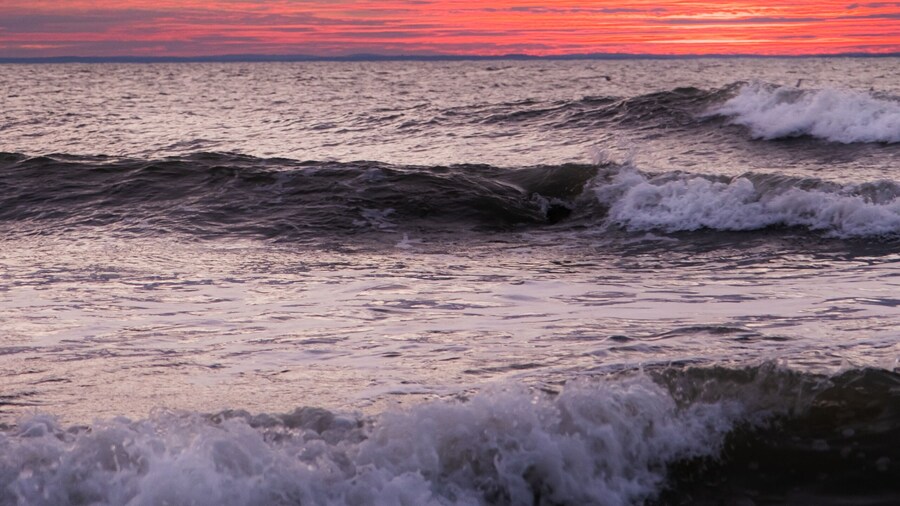 A pattern of three waves making their way to the beach and crashing on the shallow water. Each wave has a whitecap. The ocean is beneath a cloud filled sky lit by the rising sun with pink and orange.