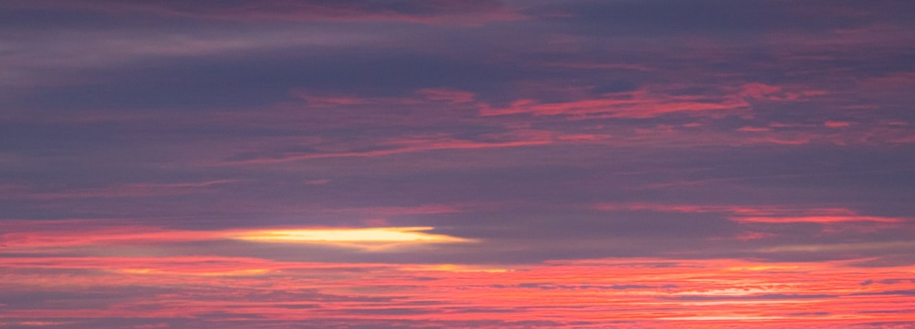A pattern of three waves making their way to the beach and crashing on the shallow water. Each wave has a whitecap. The ocean is beneath a cloud filled sky lit by the rising sun with pink and orange.