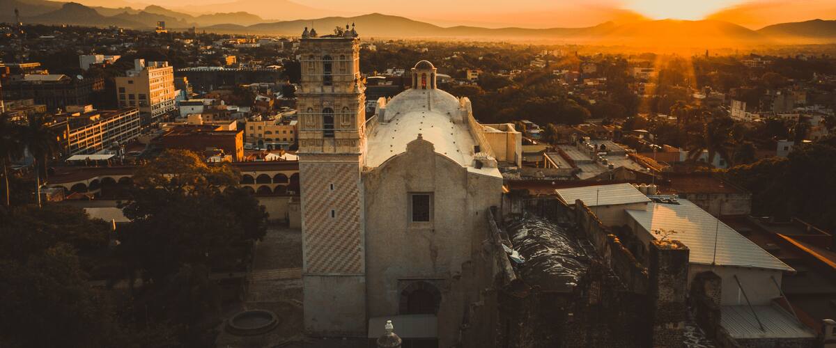 A historic church with a bell tower, surrounded by a cityscape and mountains in the background.