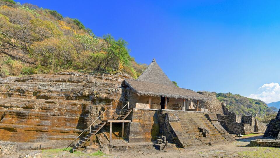A stone structure with a thatched roof, a staircase leading up to it, and a steep, rocky hillside in the background.