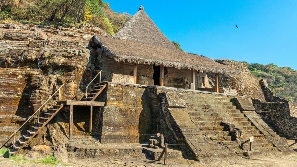 A thatched-roof structure with stone steps leading up to it, set against a rocky hillside.