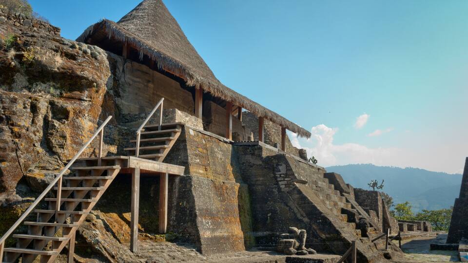 A stone structure with a thatched roof, wooden stairs, and a statue.