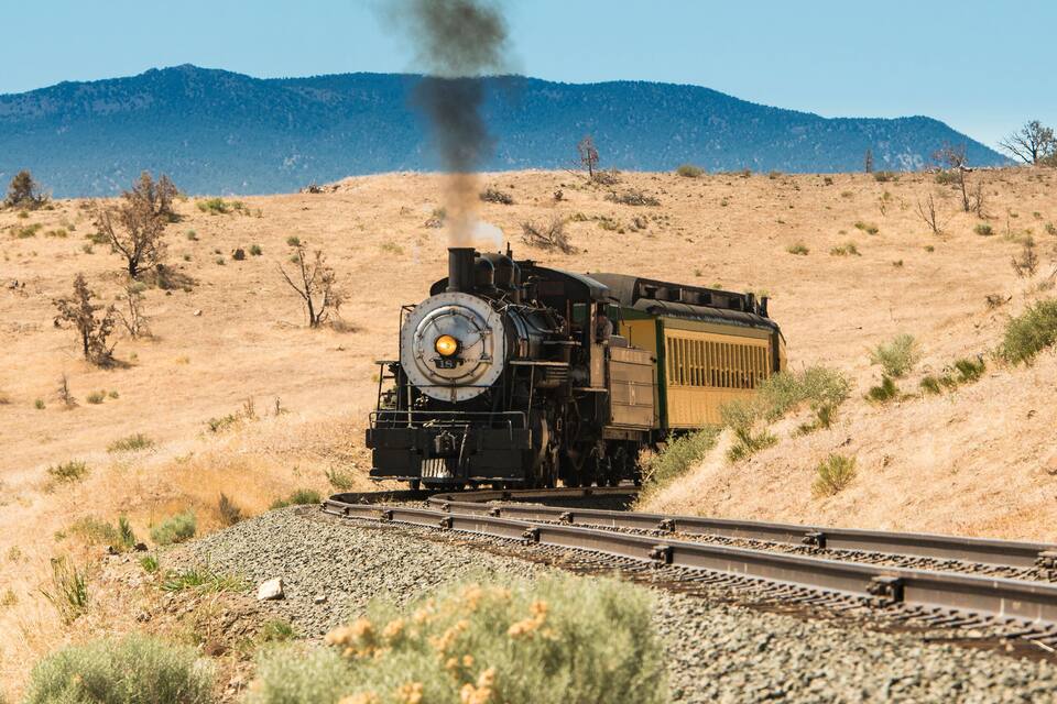 Una locomotora a vapor que allibera fum, circulant sobre vies en un paisatge sec i ondulat amb vegetació escassa.