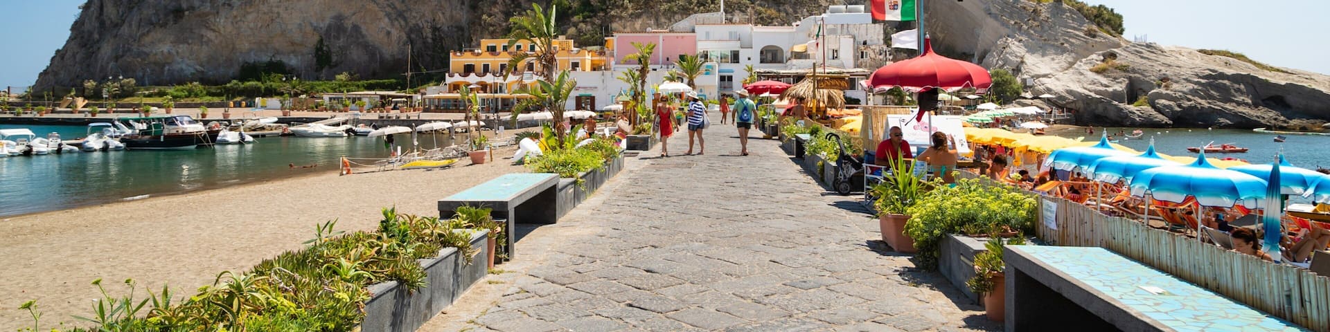 A coastal town with a prominent rocky hill, a marina with boats, and colorful umbrellas along the promenade.
