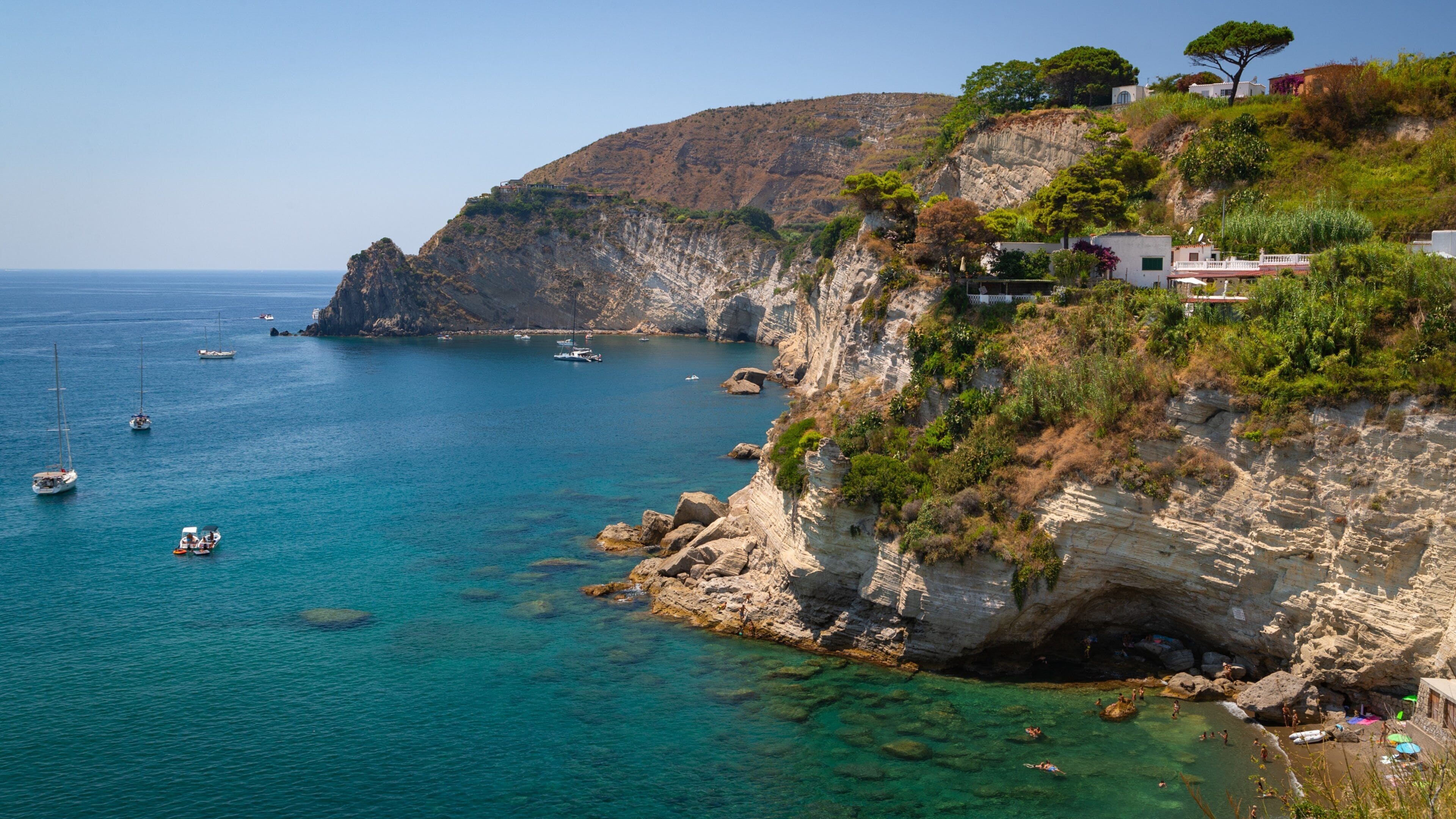 A coastal landscape with clear turquoise waters, rocky cliffs, and a few boats.