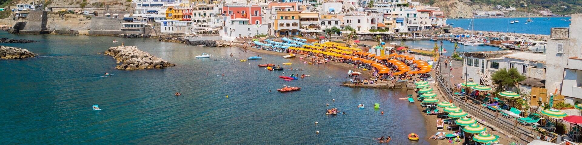 A coastal town with colorful buildings, a beach with umbrellas, and boats in the water.