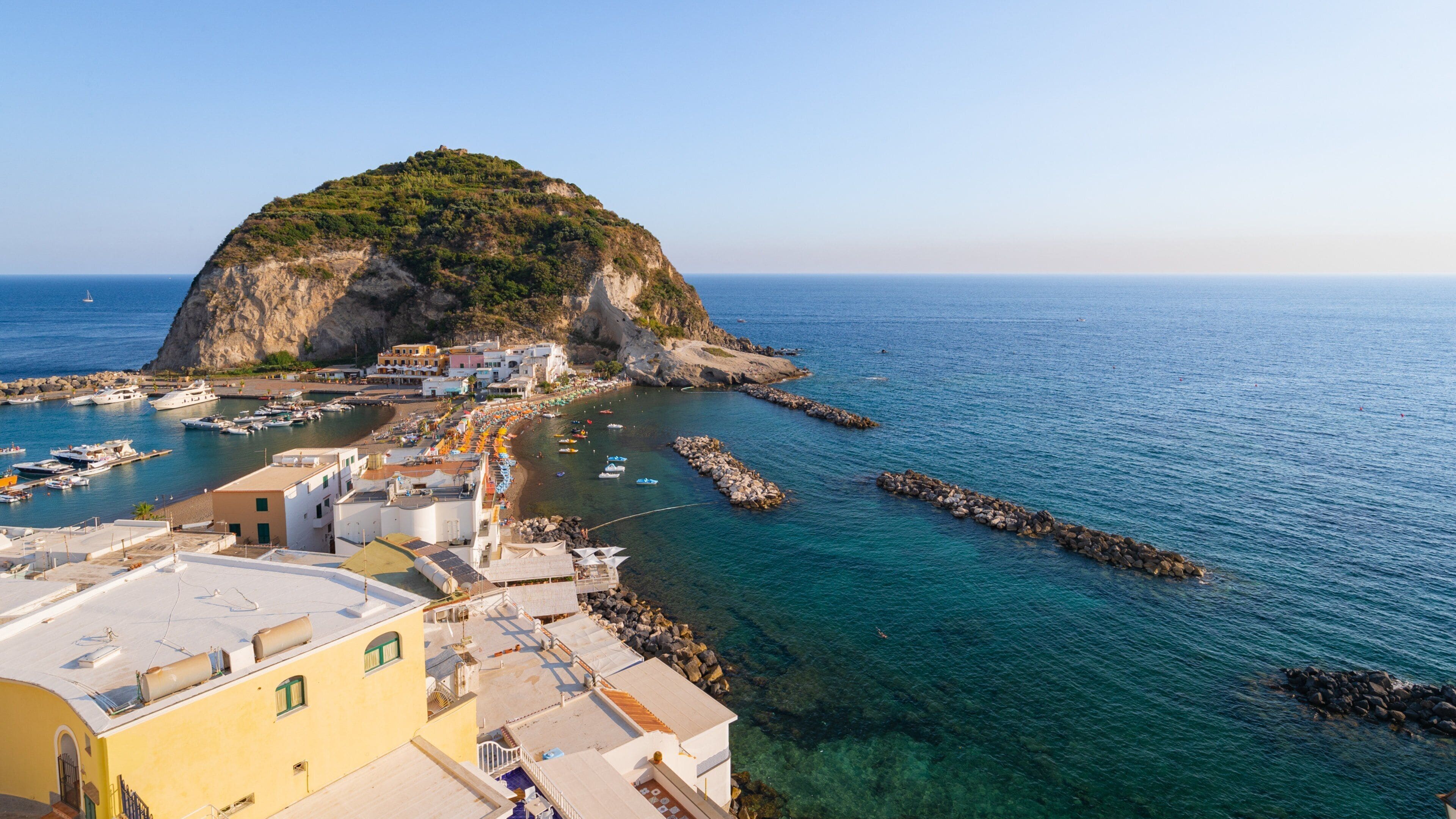 A coastal town with a prominent hill, clear blue sea, and a marina with boats.