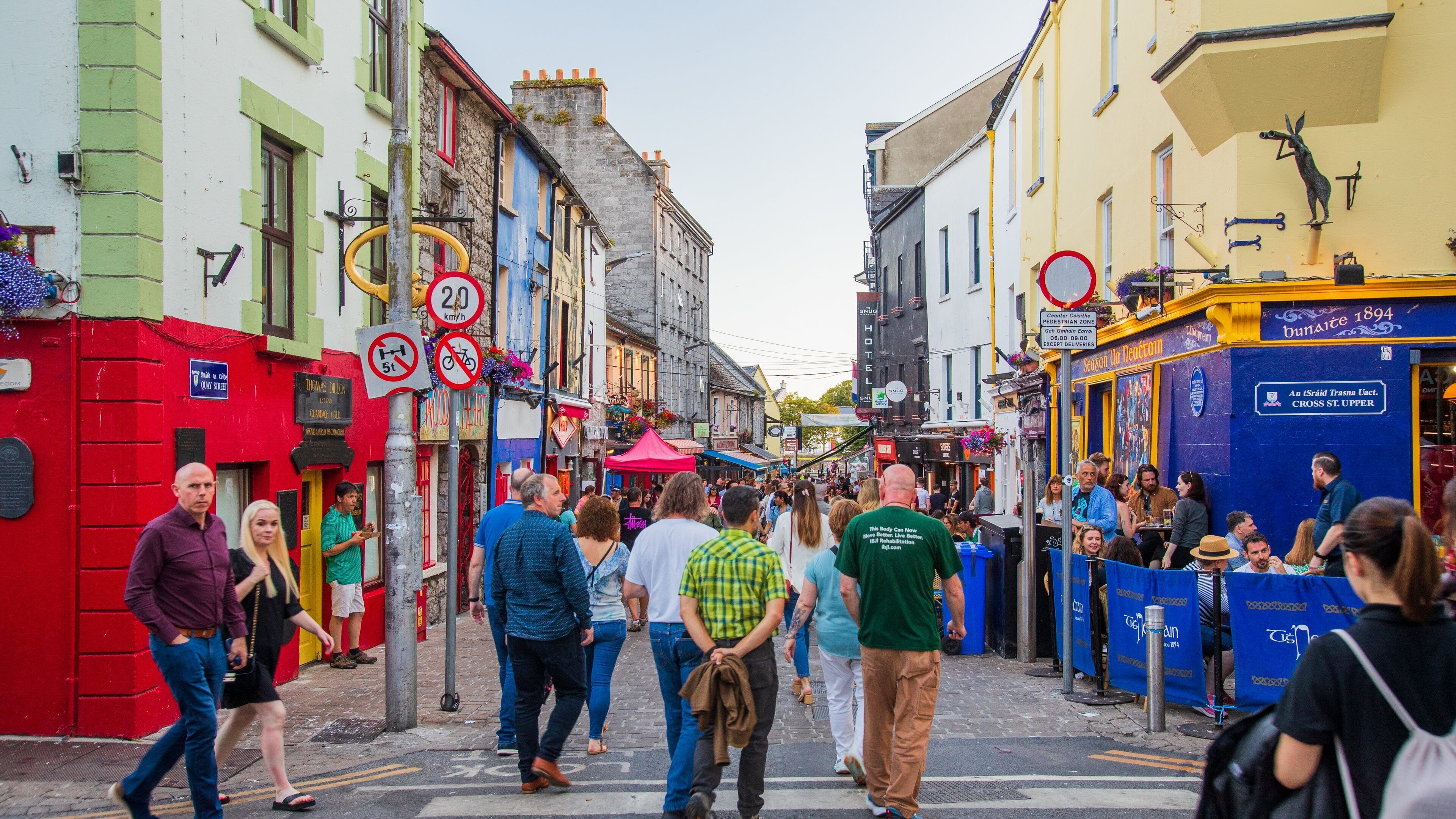 A bustling street with pedestrians, colorful buildings, and various signs including a no parking sign and a pedestrian zone sign.
