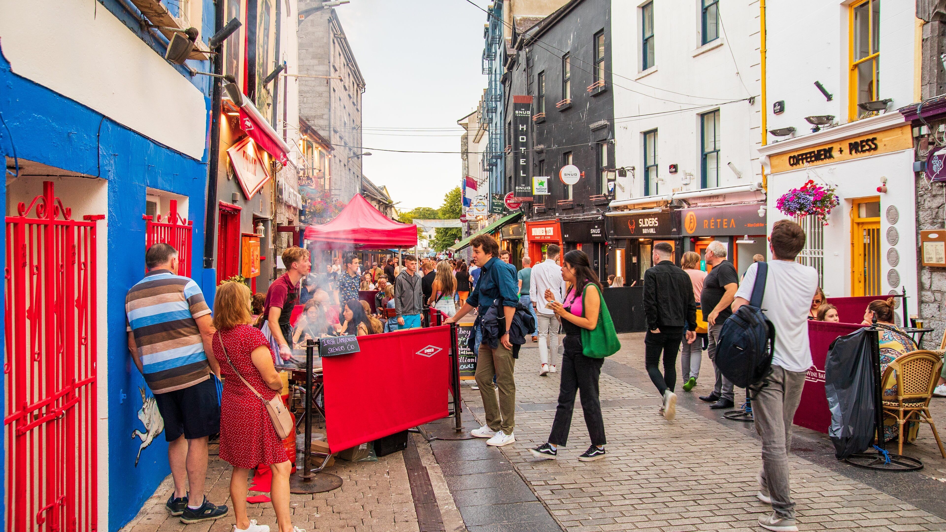 A bustling street market with people gathered around food stalls, a red barrier, and various shops and buildings.