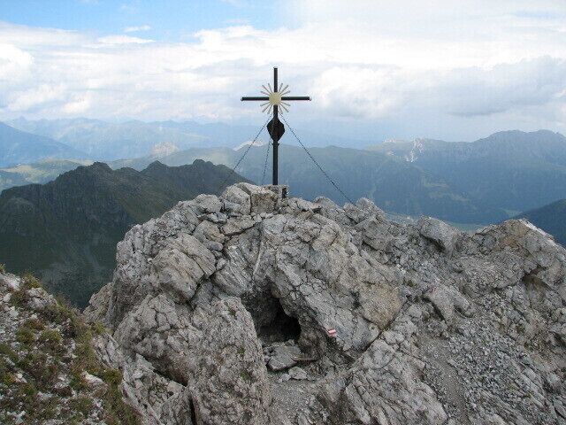 A rocky peak with a cross at the summit and a mountainous landscape in the background.