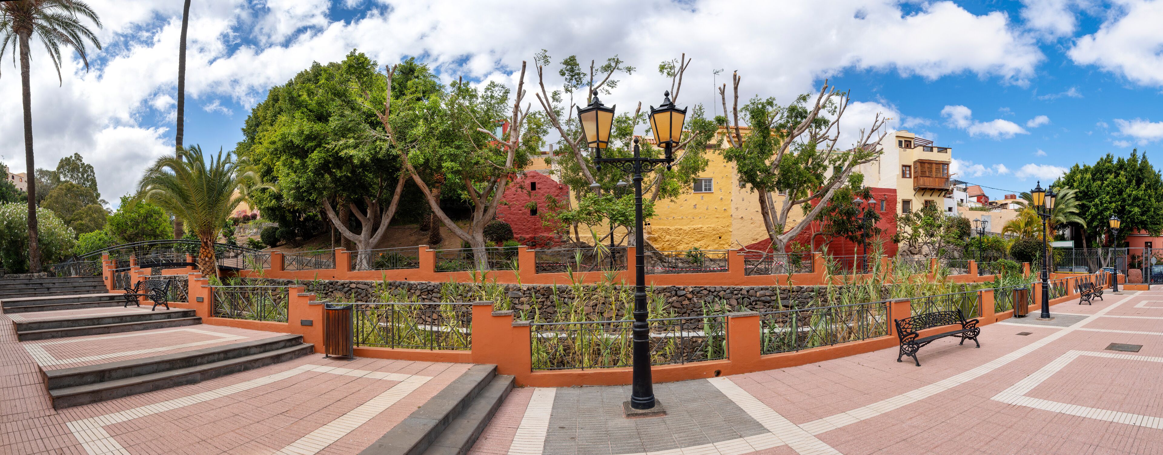 A park with a paved walkway, benches, trees, and colorful buildings in the background.
