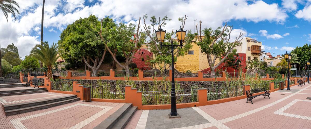A park with a paved walkway, benches, trees, and colorful buildings in the background.