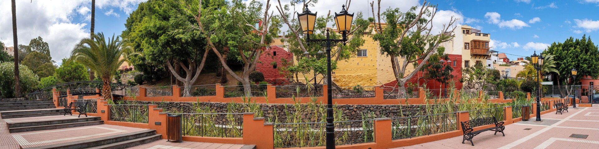 A park with a paved walkway, benches, trees, and colorful buildings in the background.