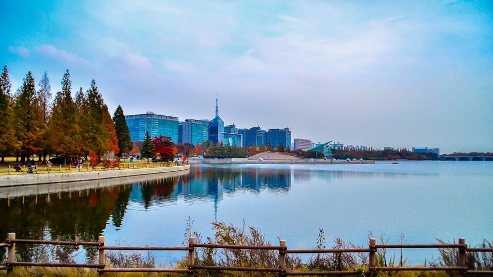 A lake with a city skyline in the background, a wooden fence in the foreground, and autumn-colored trees along the shore.