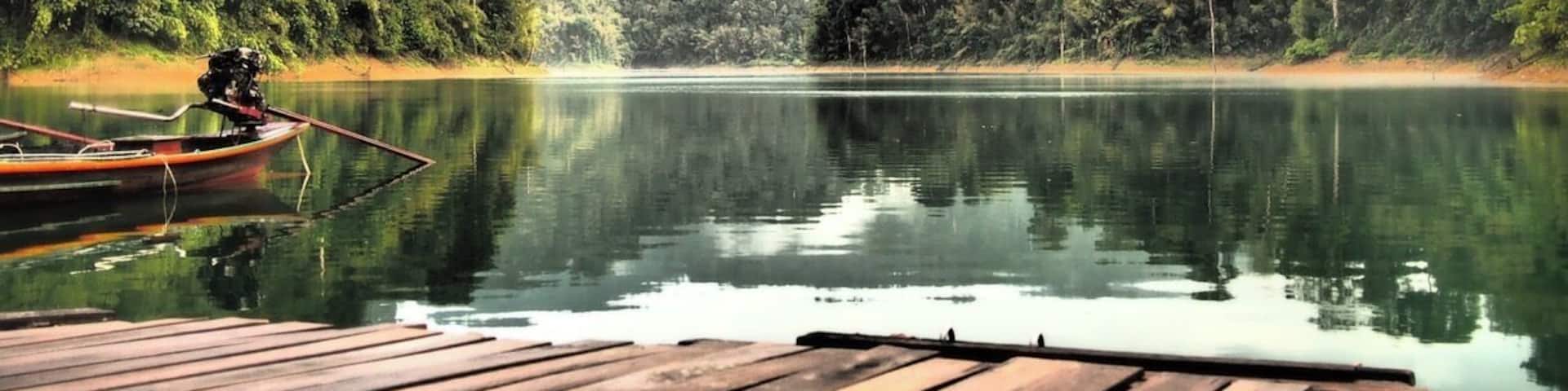 A calm lake with a wooden dock, surrounded by dense forest and misty mountains under a cloudy sky.