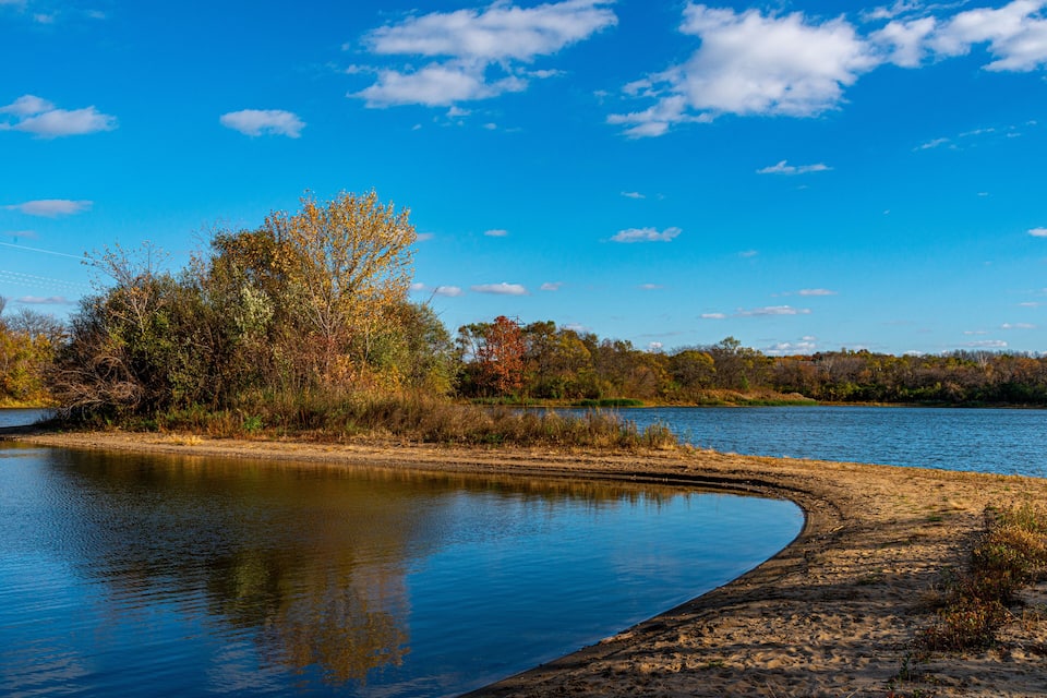Un lago tranquilo con orilla arenosa y árboles de colores otoñales bajo un cielo azul despejado.