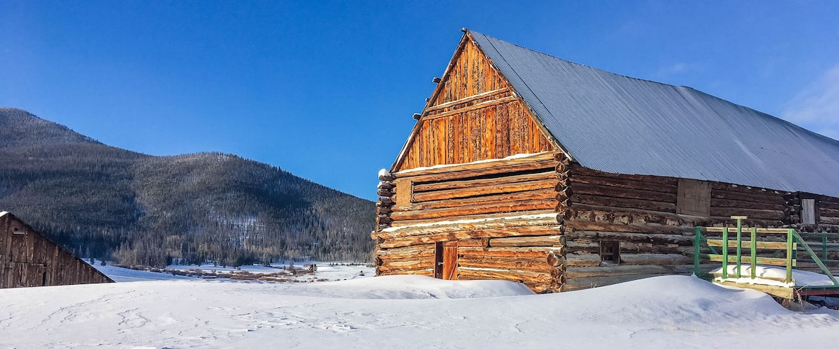 A wooden log cabin with a metal roof, surrounded by snow-covered ground and a forested mountain in the background.