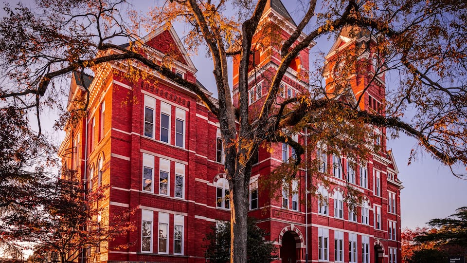 A large red brick building with arched windows and a tower, surrounded by trees with autumn leaves.