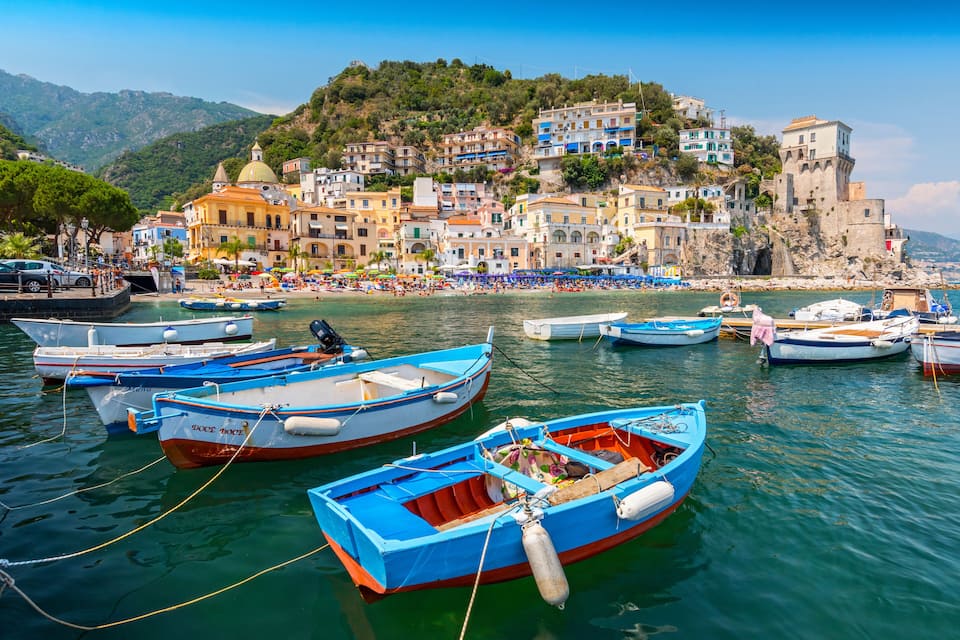 Uma vila costeira com edifícios coloridos, barcos atracados na água e montanhas ao fundo.