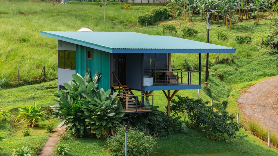 A small, single-story house with a blue roof and a balcony, surrounded by lush greenery and banana trees.