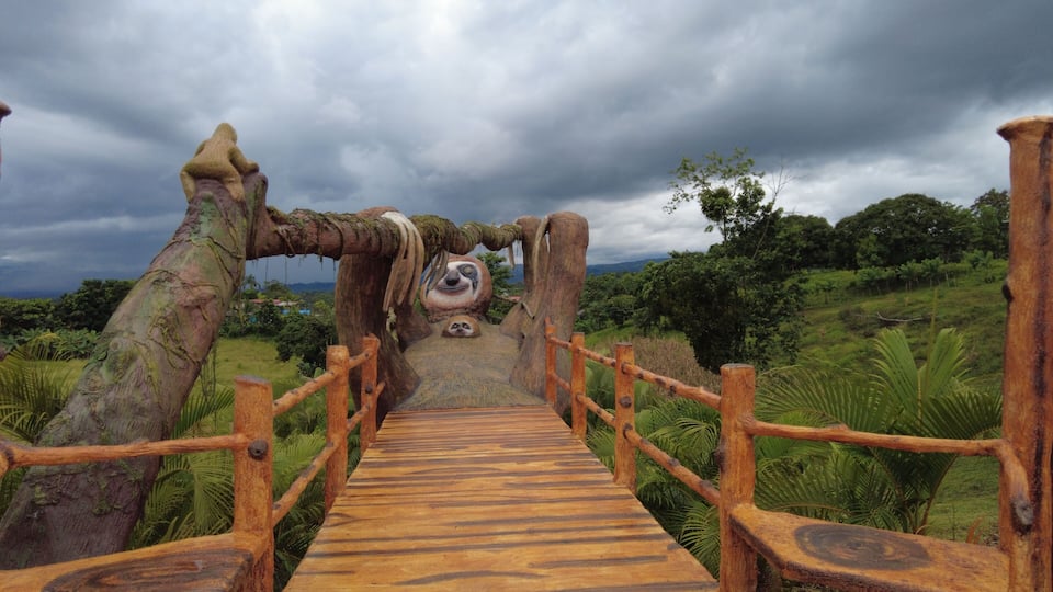 A wooden bridge with a sloth face carved into the center, surrounded by lush greenery and a cloudy sky.