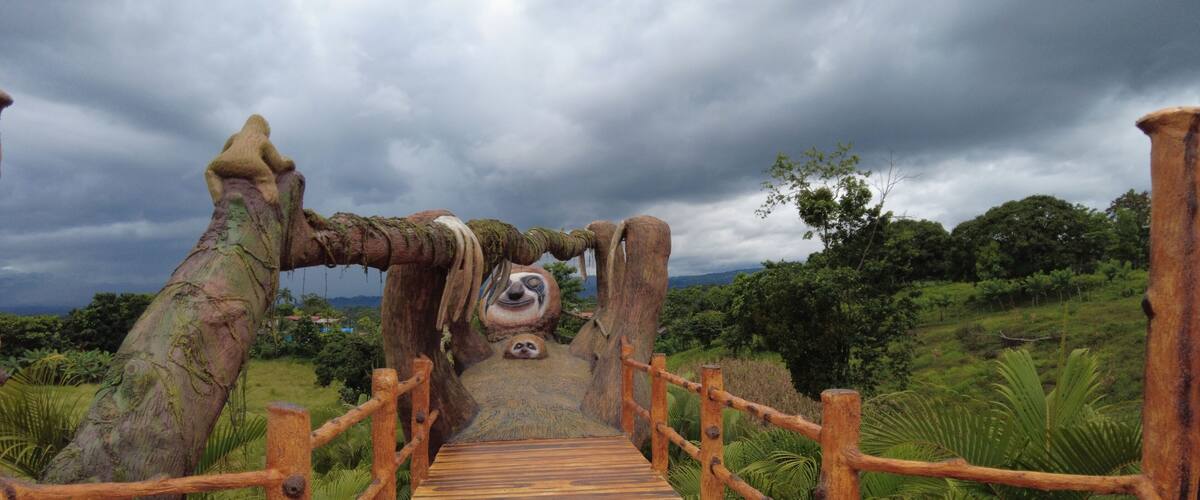 A wooden bridge with a sloth face carved into the center, surrounded by lush greenery and a cloudy sky.