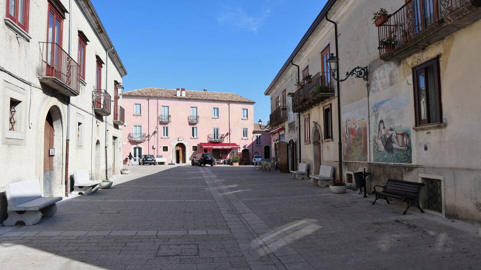 A cobblestone street lined with historic buildings, a central pink building with a red awning, and outdoor seating.