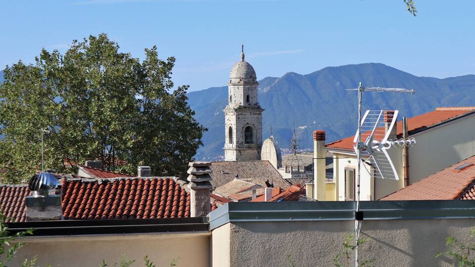 A town with red-tiled roofs, a bell tower, and mountains in the background.