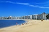 A sunny beach with people sunbathing and swimming, a lifeguard tower, and a city skyline in the background.