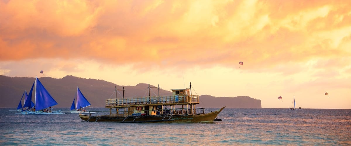 A boat with a blue sail and a wooden structure on the water, with a mountainous landscape in the background.