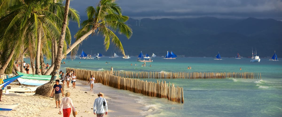 Een strand met palmbomen, een houten omheining en boten in het water.