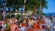 Una playa con palmeras, gente relajándose en pufs naranjas y mesas con velas.