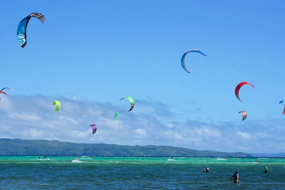 A clear blue sky with multiple colorful kites flying over turquoise waters.