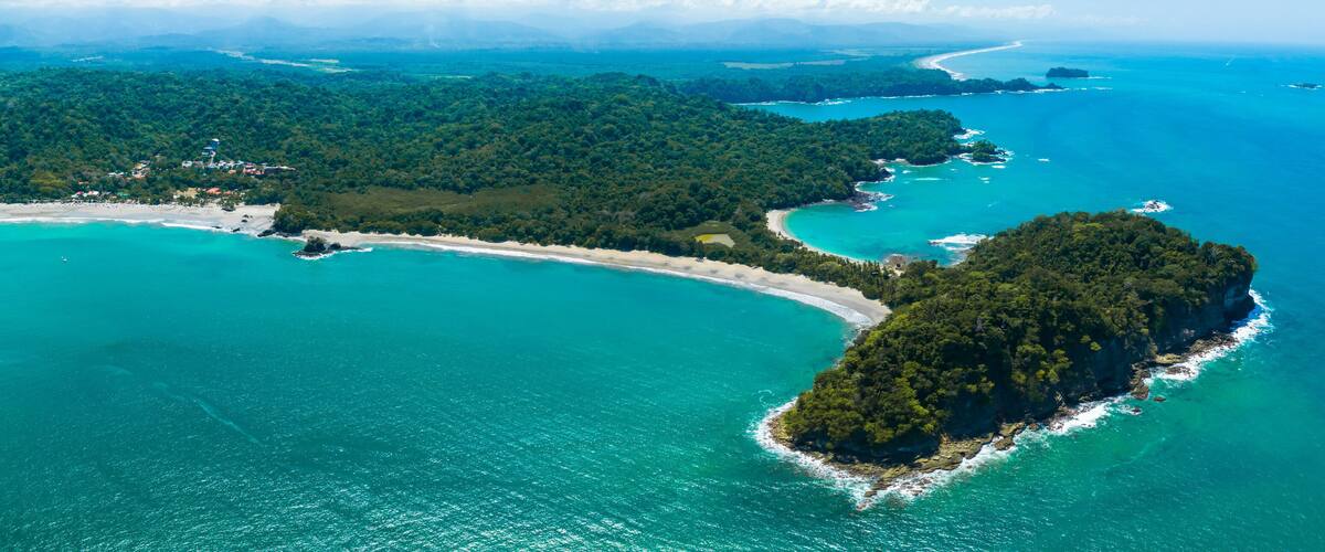 An aerial view of a coastal area with a large island surrounded by turquoise waters, a sandy beach, and dense green forests.