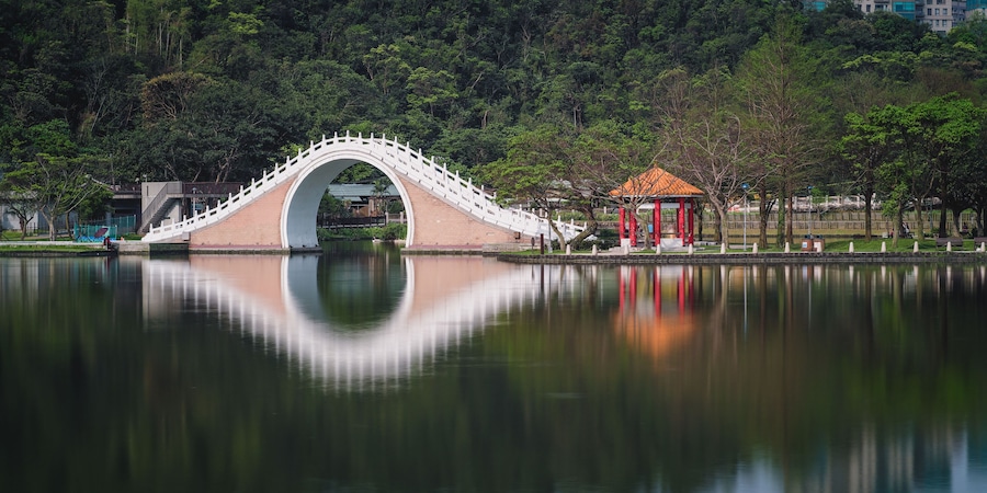 A bridge over a calm lake with a reflection, surrounded by trees and a small pavilion.