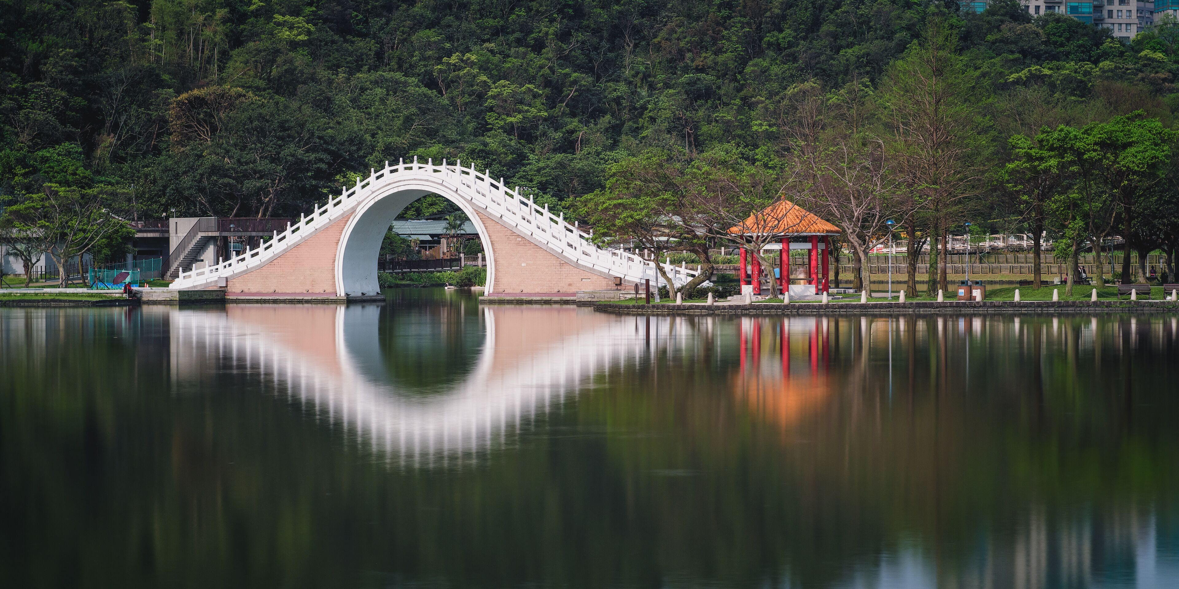 A bridge over a calm lake with a reflection, surrounded by trees and a small pavilion.