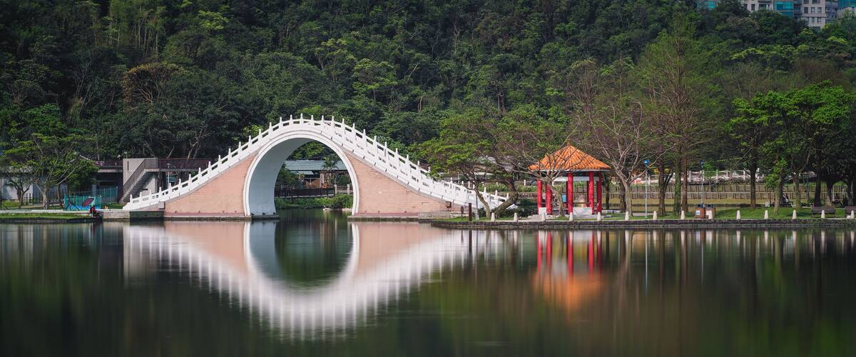 A bridge over a calm lake with a reflection, surrounded by trees and a small pavilion.