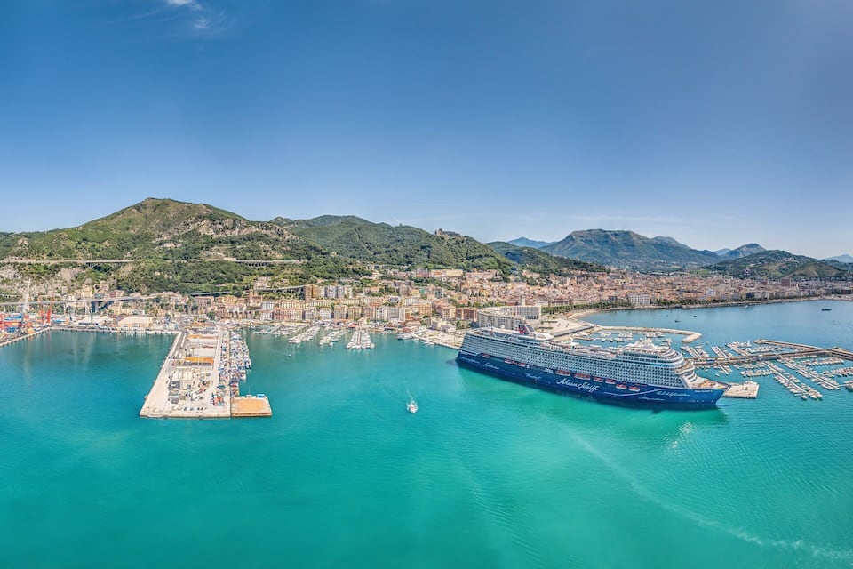 Uma cidade costeira com um grande navio de cruzeiro atracado no porto, rodeada por montanhas e um mar azul límpido.