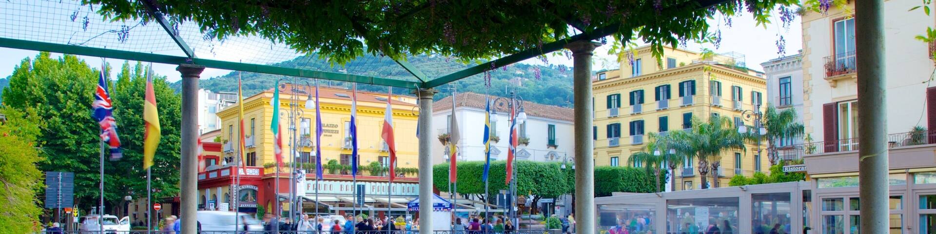 A covered outdoor café with green metal chairs and tables, overlooking a bustling street with people and buildings.
