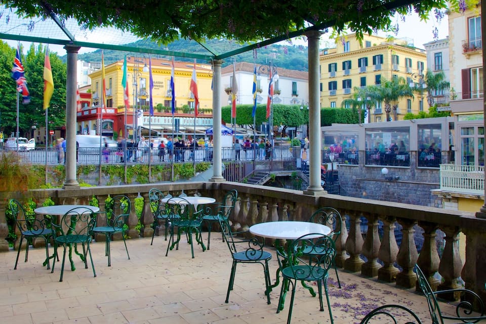 Un café al aire libre cubierto con sillas y mesas de metal verde, con vistas a una calle animada con gente y edificios.