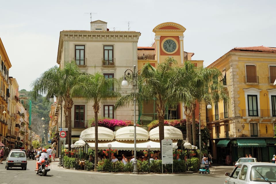 A street with palm trees, a clock tower, and outdoor cafes.