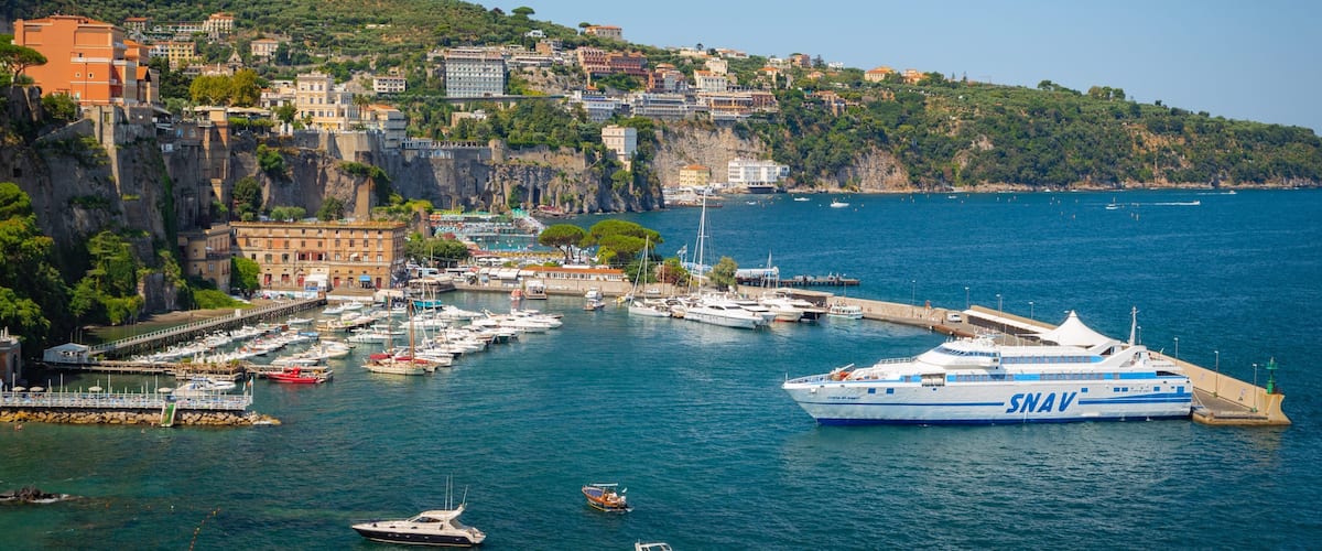 Un pueblo costero con un puerto deportivo, barcos y edificios sobre una colina.