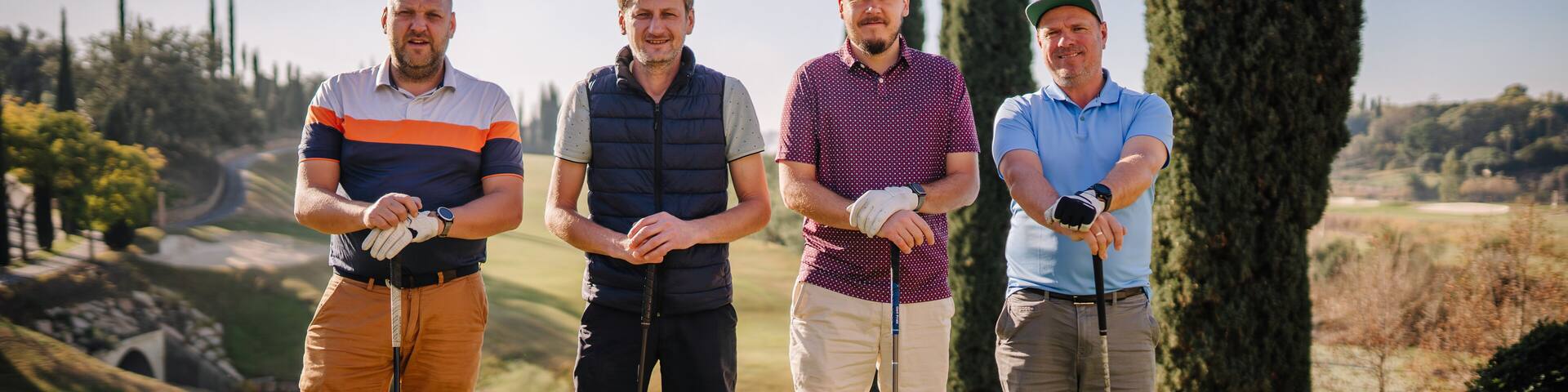 Sotogrante, Spain - January 26, 2024 - four male golfers with clubs, lined up and smiling on a golf course, with cypress trees and a clear sky in the background.