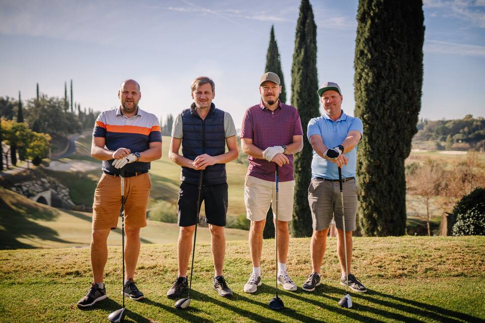 Four men standing on a golf course with a scenic background of trees and a clear sky.