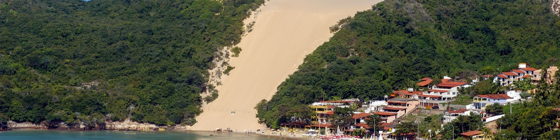 Un pueblo costero con playa, veleros y una colina con duna de arena.