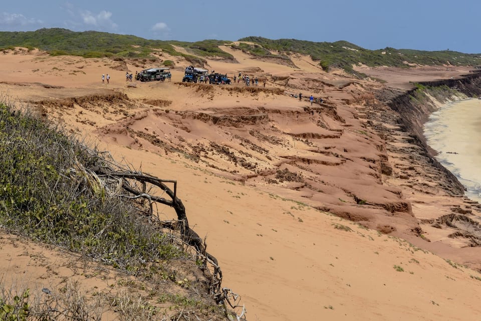 Ett kustlandskap med en sandstrand, en klippvägg och en grupp fordon på en grusväg.