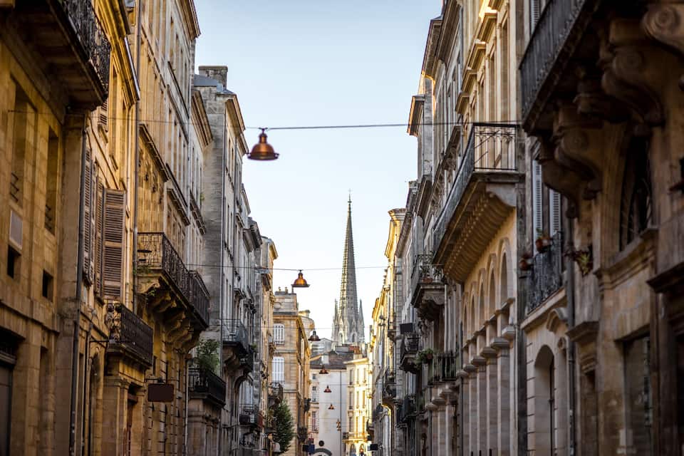 A narrow street flanked by historic buildings with ornate facades and a spire in the distance.