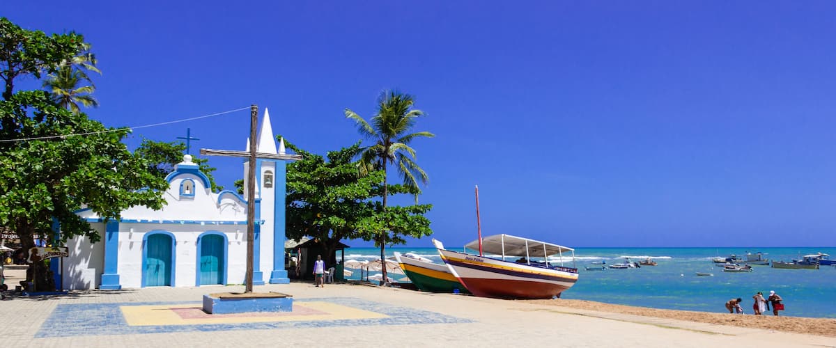 A coastal church with a blue and white facade, a large wooden cross, and a beach with boats and palm trees.