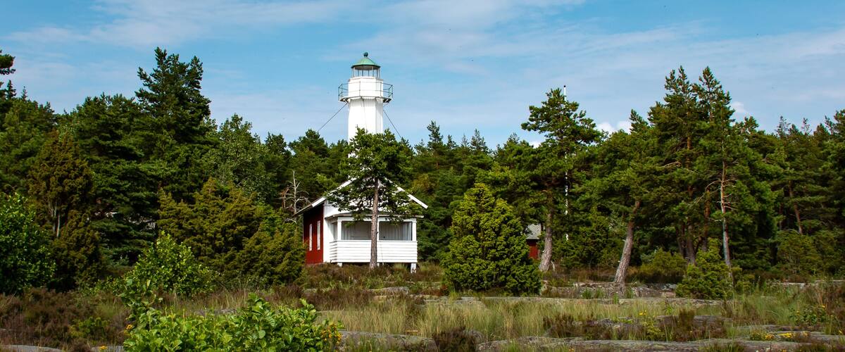 A lighthouse, a small white building, and a forested area with rocks and greenery.